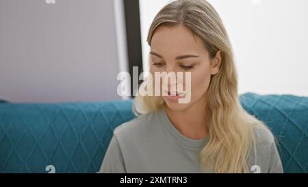 Eine nachdenkliche blonde Frau in einem lässigen Hemd sitzt drinnen, vielleicht in einem Wohnzimmer, und strahlt ein Gefühl von alltäglicher Schönheit und Ruhe aus. Stockfoto