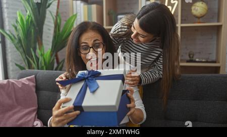 Die aufgeregte Frau erhält ein Geschenk von ihrer Tochter in einem gemütlichen Wohnzimmer, das Freude und Familienliebe ausdrückt. Stockfoto