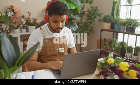 Afrikanischer Mann mit Kopfhörern, der in einem Blumenladen arbeitet, umgeben von Pflanzen. Stockfoto