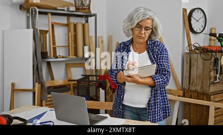 Eine reife Frau mit grauen Haaren macht sich in einem gut organisierten Tischlerstudio Notizen. Stockfoto