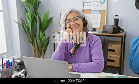 Eine lächelnde, reife Frau mit Headset arbeitet an einem Laptop in einer modernen Büroumgebung. Stockfoto