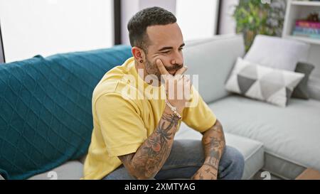 Nachdenklicher Mann mit Bart und Tattoos, der in einem modernen Wohnzimmer sitzt und ein entspanntes, stilvolles Ambiente schafft. Stockfoto