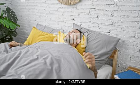 Entspannter junger Mann mit Bart und Tattoos, der in einem gemütlichen Schlafzimmer mit weißem Backsteinmauerhintergrund schläft. Stockfoto