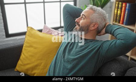 Ein reifer bärtiger Mann entspannt sich auf einer Couch in einem gemütlichen Wohnzimmer und strahlt ungezwungenen Komfort und Zufriedenheit aus. Stockfoto