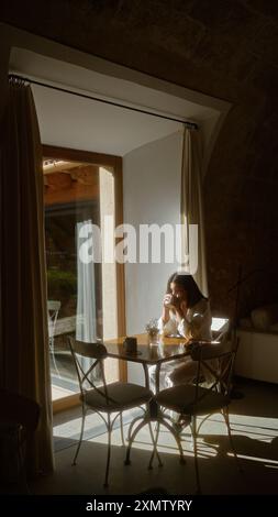 Frau genießt Kaffee in einer gemütlichen Innenumgebung mit Sonnenlicht, das durch ein großes Fenster in einem ruhigen Café hereinströmt. Stockfoto