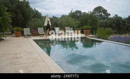 Frau, die am Swimmingpool in mallorca, spanien, vorbeiläuft, umgeben von Grün und Liegestühlen unter bewölktem Himmel, die eine ruhige Umgebung im Freien darstellen Stockfoto