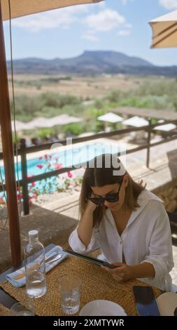 Frau liest im Freien in einer sonnigen Umgebung auf mallorca, genießt die ruhige Landschaft und den Blick auf den Pool, während sie sich ruhig mit ihrem Tablet und en beschäftigt Stockfoto