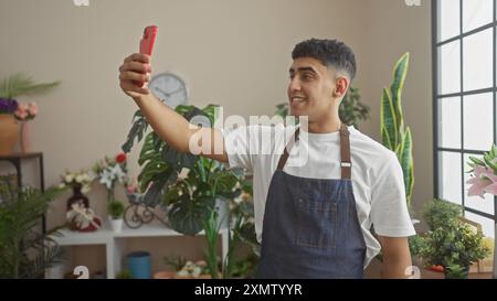 Ein lächelnder junger Mann in einer Schürze, der ein Selfie in einem Blumenladen macht, geschmückt mit verschiedenen Pflanzen und Blumen. Stockfoto