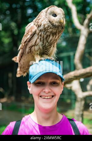 Porträt einer lachenden 36-jährigen russischen Frau mit Ural-Eulen (Strix uralensis) auf dem Kopf sitzendem Vogel Stockfoto