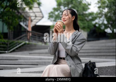 Eine attraktive, entspannte asiatische Geschäftsfrau genießt ihren Kaffee, während sie auf der Treppe im Freien sitzt, ihre Augen geschlossen hält und nach Kaffee riecht Stockfoto