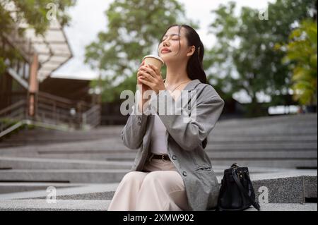 Eine attraktive, entspannte asiatische Geschäftsfrau genießt ihren Kaffee, während sie auf der Treppe im Freien sitzt, ihre Augen geschlossen hält und nach Kaffee riecht Stockfoto