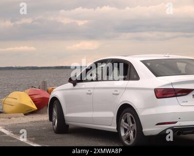 Weißes Auto parkte in der Nähe des Wassers mit einem kleinen Hund, der vom Fahrersitz auf die Rückseite des Autos blickte. Rote und gelbe Kajaks auf Sand. Stockfoto