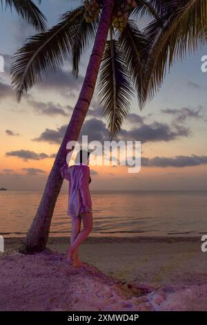 Eine Frau in einem hellrosa Hemd steht an einem Sandstrand, lehnt sich an eine Palme und beobachtet den Sonnenuntergang über dem Meer. Stockfoto