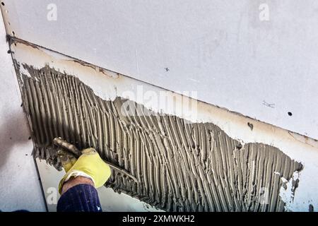 Die Fliesenmaschine des Bauarbeiters trägt mit einer gekerbten Kelle Klebermörtel auf die Wand auf. Stockfoto