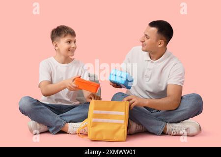 Der kleine Junge und sein Vater packen Schulbrotdosen in einen Beutel auf rosa Hintergrund Stockfoto