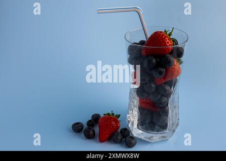 Ein bis zum Rand gefülltes Glas mit einer bunten Auswahl frischer, reifer Beeren, begleitet von einem Strohhalm. Die Beeren sind ordentlich angeordnet, wodurch ein Visu entsteht Stockfoto