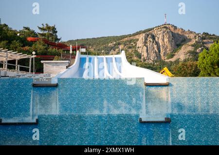 Eine große blaue Wasserrutsche mit weißem Dach befindet sich vor einem Pool. Im Hintergrund befindet sich ein Berg mit einer hohen Struktur auf dem Gipfel. Familienurlaub Stockfoto