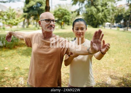 Älterer Mann und asiatische Frau, die Tai Chi in einem friedlichen Park praktizieren, umgeben von Grün, konzentrieren sich auf präzise Bewegungen mit entspannten, ruhigen Ausdrücken. Stockfoto