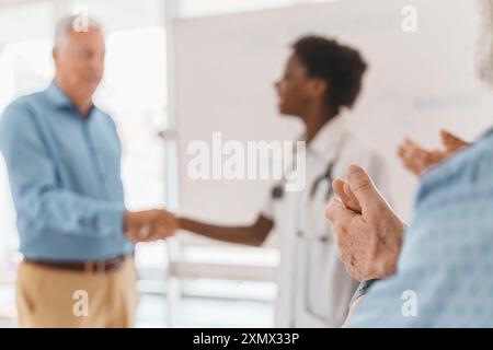 Ältere Hände applaudieren, als afroamerikanische Frau mit einem älteren Mann im Hintergrund die Hand schüttelt. Szene zeigt akademische oder berufliche Leistungen, mento Stockfoto