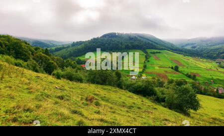 Berglandschaft im Herbst. Grasbewachsene Wiese auf einem Hügel. Dorf im fernen Tal. Ländliche Landschaft. Regnerisches Wetter mit bewölktem Himmel Stockfoto