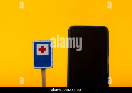 Ein hölzernes Krankenhaus-Warnschild in Verbindung mit einem Smartphone auf gelbem Hintergrund, das den Kontrast zwischen alten und neuen Kommunikationsmitteln unterstreicht Stockfoto