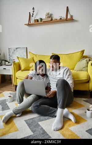 Ein liebevolles Paar sitzt auf dem Boden vor einer gelben Couch und stöbert auf einem Laptop. Stockfoto