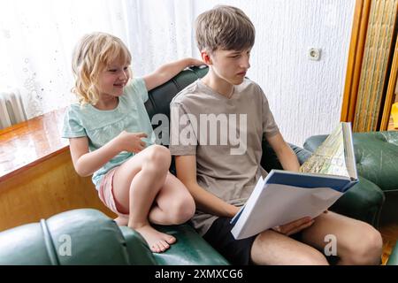 Ein kleiner Junge sitzt auf einer grünen Couch in seinem Haus und liest seiner kleinen Schwester ein Buch vor, die neben ihm sitzt und mit einem Lächeln zu ihm aufblickt. Stockfoto