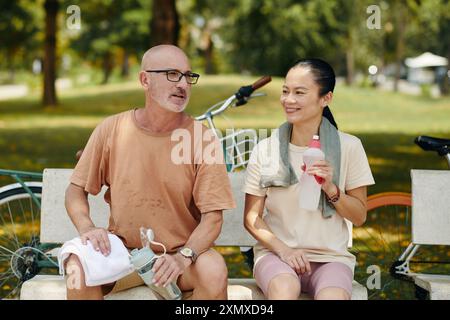 Lächelnder älterer kaukasischer Mann und asiatische Frau sitzen auf einer Bank im Park, mit Fahrrädern im Hintergrund und genießen Gespräche, während sie mit drin hydratisieren Stockfoto