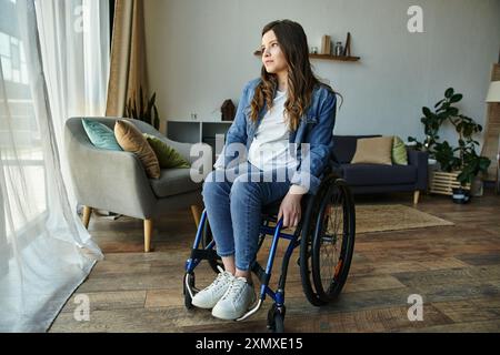 Eine junge Frau im Rollstuhl sitzt in ihrer modernen Wohnung und schaut aus dem Fenster. Stockfoto