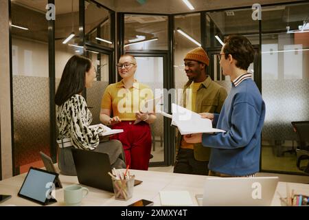 Gruppe von vier ethnisch unterschiedlichen jungen Kollegen, die Dokumente in einem modernen Büro halten und Gespräche führen Stockfoto