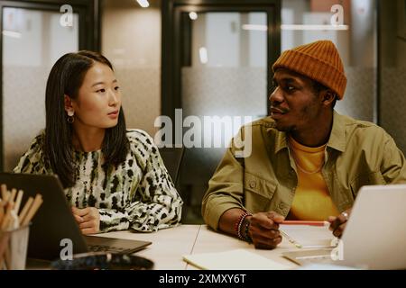 Junger afroamerikanischer Designer und asiatischer Manager arbeiten gemeinsam an einem neuen Projekt, während er am Schreibtisch in einem modernen Büro sitzt Stockfoto