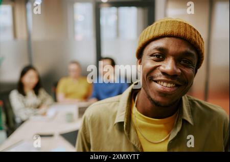 Mittelgroßes Nahaufnahme-Porträt eines glücklichen jungen afroamerikanischen Mannes, der legere Kleidung trägt, im Büro steht und in die Kamera lächelt Stockfoto