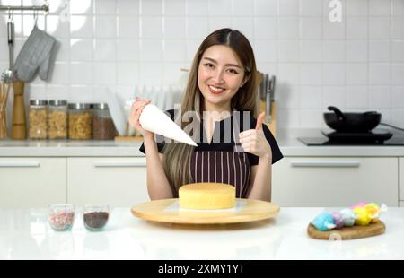 Die junge Frau in einer Schürze hebt die Daumen hoch, während sie den Saugbeutel hält. Bereiten Sie sich darauf vor, die Glasur auf den Kuchen zu gießen, der auf einem Holzdrehtisch oder platziert wird Stockfoto