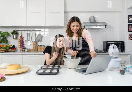 Zwei junge Frauen auf einer Schürze bereiten in der Küche Nachtisch zu, während sie auf dem Laptop im Online-Rezeptbuch nachschauen. Stockfoto