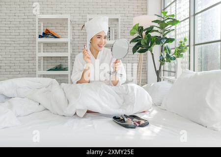 Junge asiatische Frau mit einem Lächeln, auf einem Bett sitzen und Make-up auftragen, einen Spiegel halten, einen Bademantel und ein Handtuch auf dem Kopf tragen. Stockfoto