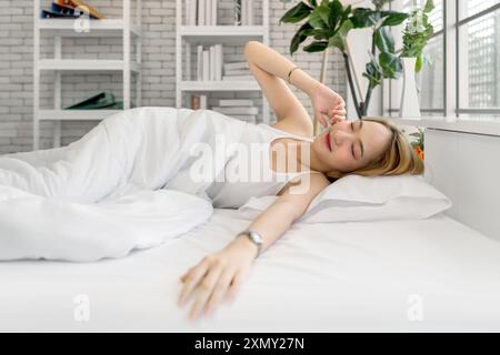 Junge asiatische Frau in weißem Unterhemd, die in einem hellen, gemütlichen Zimmer mit Kissen und Bettdecke auf dem Bett liegt. Seitenansicht Stockfoto