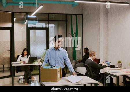 Junger birassischer Mann, der am Schreibtisch im Büro eines führenden Unternehmens steht und sich auf den ersten Arbeitstag vorbereitet, Dokumente und anderes aus dem Karton nimmt Stockfoto