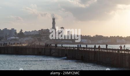 Alexandria, Ägypten - 19. Dezember 2018: Küstenlandschaft mit Menschen auf Fußgängerbrücken unter farbenfroher bewölktem Himmel, Montazah Palast im Hintergrund Stockfoto