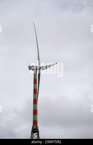 Hoch vor dem Hintergrund eines bewölkten Himmels erwartet Sie eine hoch aufragende Windenergieanlage, deren drei Rotorblätter bereit sind, die Kraft des Windes einzufangen. Verziert mit Stockfoto