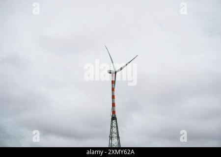 Die hoch aufragende Windturbine steht vor einem bewölkten Himmel, und ihre drei Rotorblätter sind so positioniert, dass sie die Kraft des Windes einfangen. Die orangefarbene und weiße Stk Stockfoto