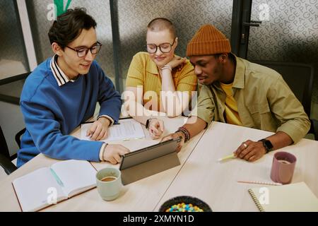 Hochwinkelansicht ethnisch vielfältiger junger Männer und Frauen, die auf den digitalen Tablet-Bildschirm schauen, während sie am Schreibtisch im Büro sitzen Stockfoto