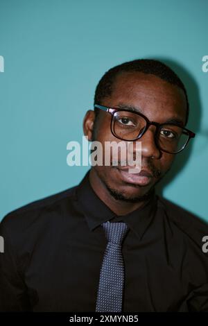 Vertikaler Blitz Porträt eines jungen afroamerikanischen männlichen Mitarbeiters mit Brille und Blick auf die Kamera, blauer Hintergrund Stockfoto