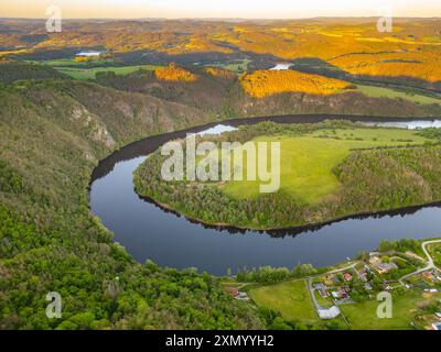 Ein malerischer Blick aus der Luft auf die Moldau-Biegung, Solenice Hufeisen, durch die tschechische Landschaft bei Sonnenuntergang. Üppige grüne Bäume und Felder rund um den Fluss, während die Sonne ein goldenes Leuchten über die Landschaft wirft. Stockfoto