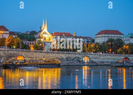 Die Moldau fließt am beleuchteten Emmaus-Kloster in Prag, Tschechien, vorbei. Das Wasser reflektiert die Lichter der Stadt und schafft eine wunderschöne Szene. Ein Boot liegt nahe dem Fluss an. Stockfoto