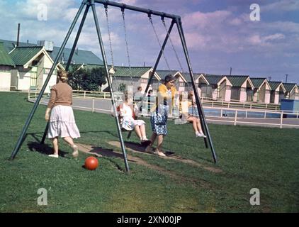 1960er Jahre, historisch, eine Mutter mit ihren beiden kleinen Kindern auf Schaukeln auf dem Grasgelände außerhalb der einstöckigen Holzchalets in einem britischen Ferienlager in England, Großbritannien. Stockfoto