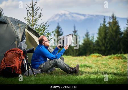 Der Mann sitzt neben dem Touristenzelt und hält ein tragbares Solarmodul-Ladegerät, um Sonnenlicht einzufangen und die Powerbank aufzuladen. Großer oranger Rucksack, Kamera auf Stativ. Schneebedeckte Berge unter teilweise bewölktem Himmel. Stockfoto