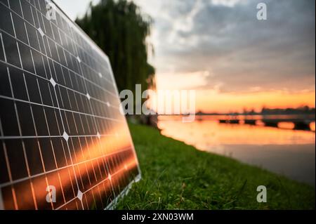 Sonnenkollektor, beleuchtet von der untergehenden Sonne, steht am grasbewachsenen Seeufer, mit malerischem Sonnenuntergang und Bäumen im Hintergrund. Fusion erneuerbarer Energietechnologien mit ruhiger Naturlandschaft. Stockfoto