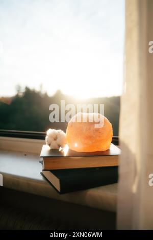 Eine Himalaya-Salzlampe strahlt ein warmes Leuchten auf einen Stapel Bücher aus, die auf einer Fensterbank platziert sind. Die Sonne geht im Hintergrund unter und wirft ein sanftes Licht. Die Atmosphäre Stockfoto