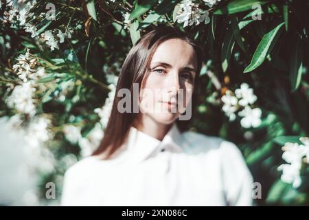 Ein wahres Neigeporträt einer Frau in einem weißen Hemd steht vor üppig grünem Laub und weißen Blumen. Das Foto unterstreicht ihre Ruhe Stockfoto