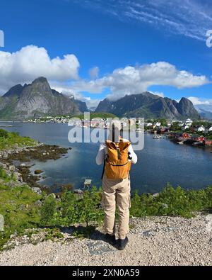 Ein Wanderer bestaunt die atemberaubenden Fjorde der Lofoten Islands, umgeben von dramatischen Gipfeln und lebhaften Dörfern unter einem hellblauen Himmel. Reine Lofoten Norwegen Stockfoto